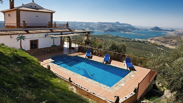 A villa with a swimming pool, blue lounge chairs, and a view of the surrounding landscape.