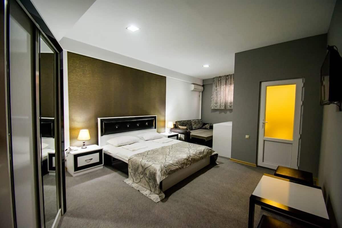 A hotel room with a bed, bedside tables, a desk, a chair, a lamp, and a door with a light.