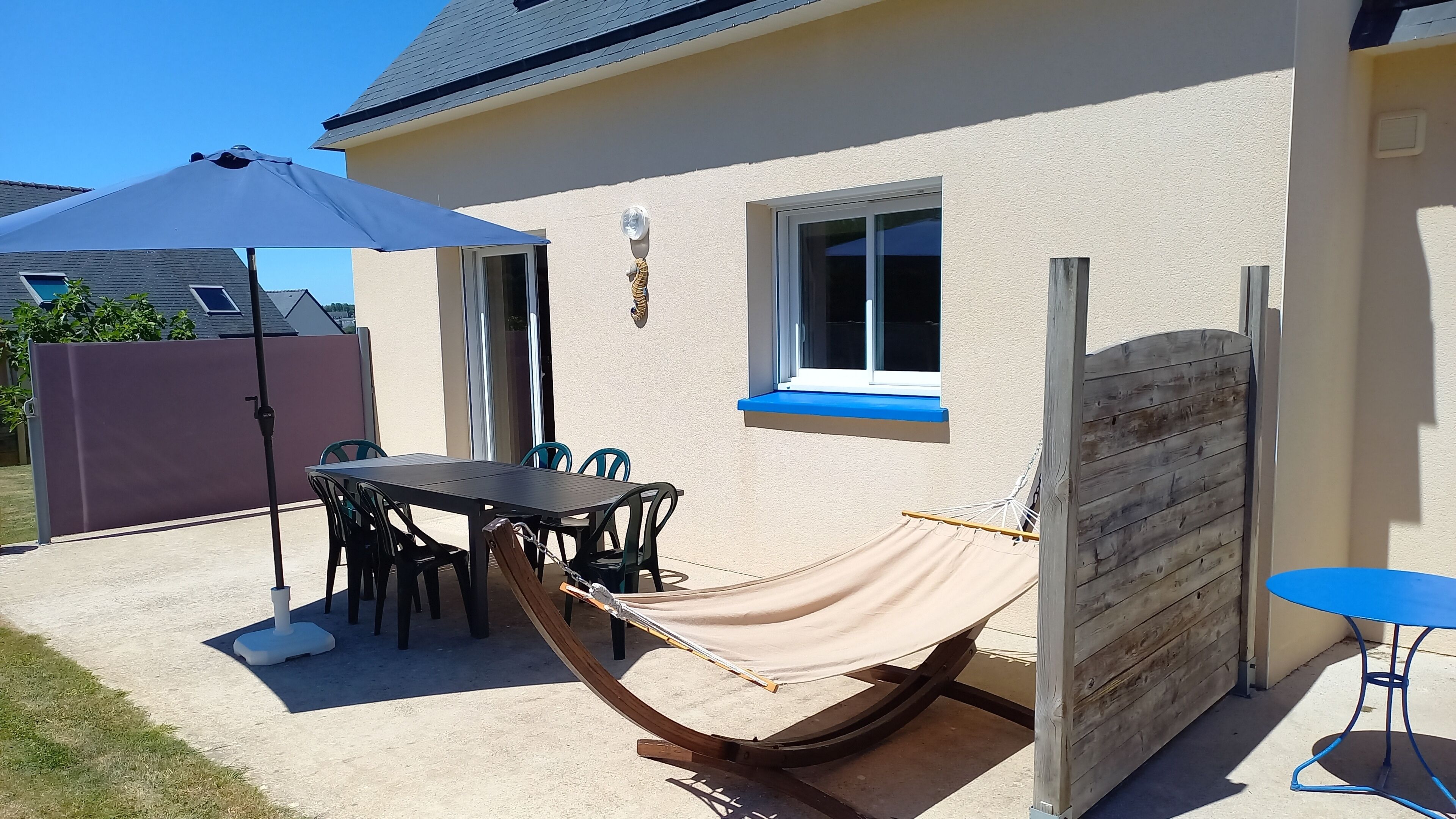Een terras met een tafel, stoelen, een hangmat, een blauwe tafel en een houten omheining.