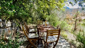 A rustic outdoor dining area with wooden tables and chairs, surrounded by natural vegetation and a stone wall.