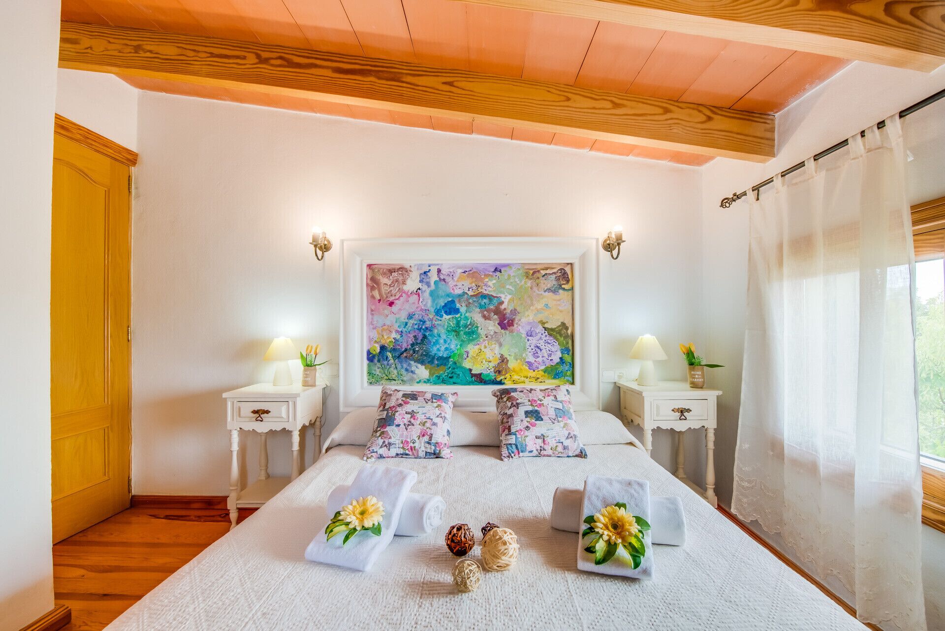 Une chambre à coucher avec un grand lit, deux tables de chevet, un tableau au mur et une fenêtre avec des rideaux.