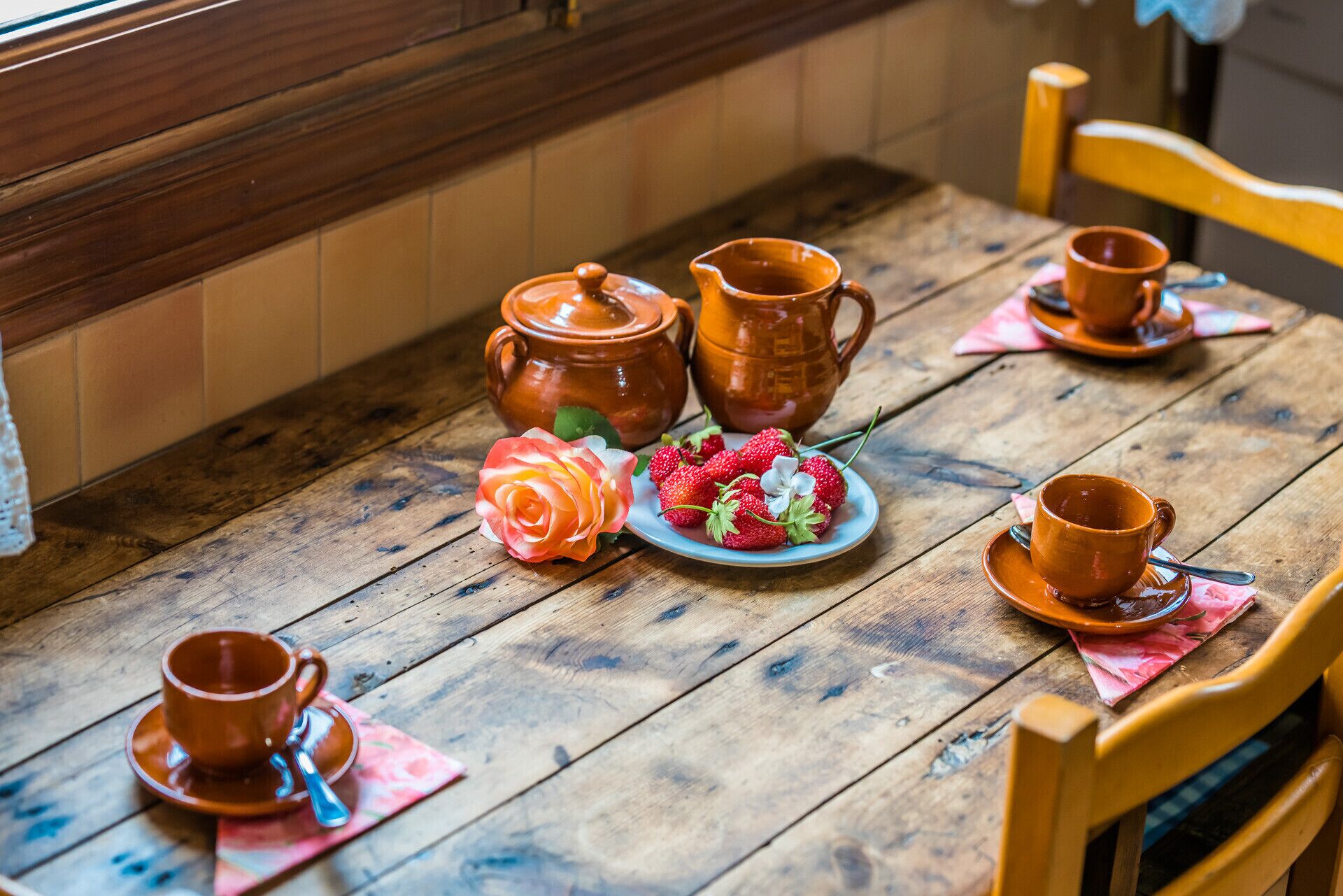 A wooden table with ceramic tea sets, a plate of strawberries, and a decorative rose.