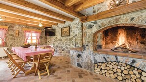 A rustic stone fireplace with a fire, wooden beams, a stone wall, and a table with checkered tablecloths and wooden chairs. - 4 bedroom pet friendly home in Perusic (Perusic)