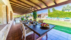 Un área de comedor al aire libre con una mesa larga dispuesta para una comida, rodeada de sillas de mimbre y con vistas a una piscina y jardín.