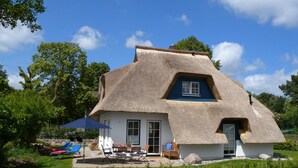 Ein reetgedecktes Haus mit blauem Fenster, einer Terrasse mit Gartenmöbeln und einem Garten.
