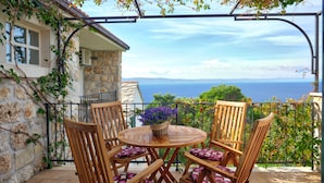 A wooden table with chairs and a potted plant on a balcony with a stone wall and a view of the sea.