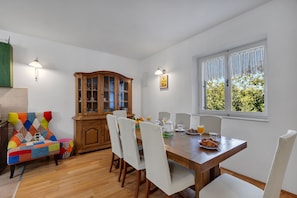A dining area with a wooden table, white chairs, and a colorful armchair. A wooden cabinet and a window with curtains are also visible. - Villa with infinity pool, teracce and sea view, bbq, internet, two bedrooms (Tucepi)