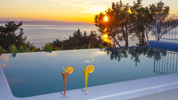 An infinity pool with two cocktails, sunset view, and a metal railing.