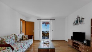 A living room with a sofa, a glass coffee table, a TV on a wooden stand, and a view through a door.