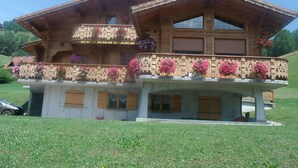 A two-story wooden chalet with balconies adorned with flowers, set in a grassy area with a car parked nearby. - Ground floor apartment for rent in Bernex for 5 people (Bernex)