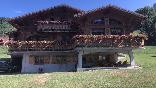 A wooden chalet with a balcony adorned with flowers, a covered outdoor seating area, and a car parked in front. - Ground floor apartment for rent in Bernex for 5 people (Bernex)