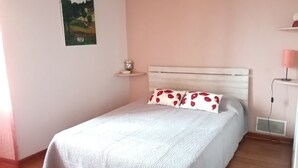 Un lit bien fait, recouvert d’une couvre-lit blanche et agrémenté de deux oreillers ornés de motifs floraux rouges, une petite table de chevet avec un vase, et un tableau représentant un paysage accroché au mur.
