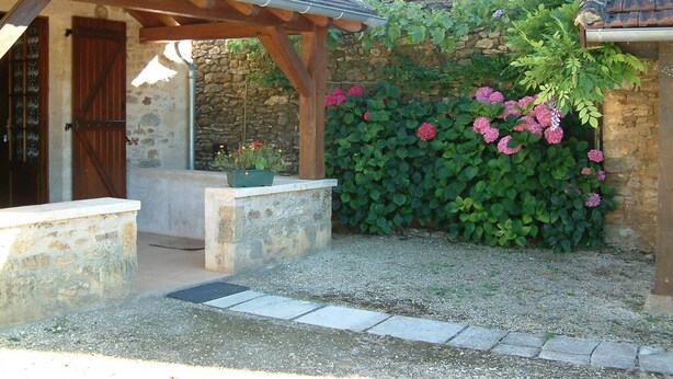 Un bâtiment en pierre agrémenté d’une pergola en bois, un mur en pierre recouvert de plantes grimpantes et un chemin de gravier.