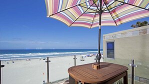 Une table et des chaises en bois, placées sous un parasol à rayures, sur un balcon donnant sur une plage, avec un ciel dégagé.