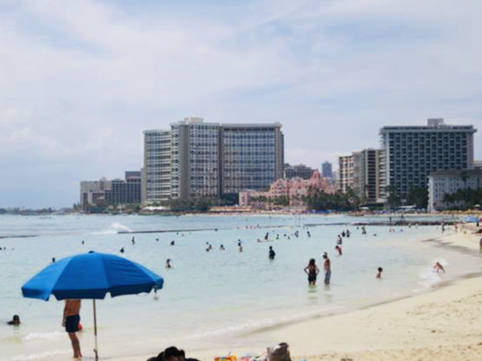 A beach with people swimming and standing in the water, a blue umbrella, and tall buildings in the background.