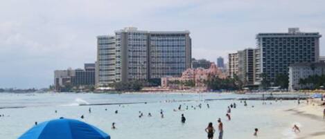 A beach with people swimming and standing in the water, a blue umbrella, and tall buildings in the background.
