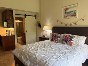 A bedroom with a bed, a nightstand, a lamp, a picture on the wall, and a door leading to a bathroom.