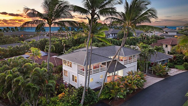 Ein zweistöckiges Haus mit Balkon, umgeben von üppiger Vegetation und Palmen, mit Blick auf das Meer bei Sonnenuntergang.