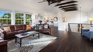 Ein geräumiger Wohnbereich mit Deckenventilator, einem braunen Ledersofa, einem Couchtisch und Blick auf die Küche und den Essbereich.
