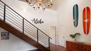 Eine Treppe mit Holzgeländer und Metallspindeln, eine Wand mit dem Schriftzug "Aloha" sowie zwei Surfbrettern und ein Holzkonsolentisch mit einer Pflanze.