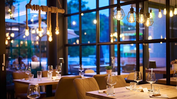 A restaurant interior with tables set for dining, hanging light fixtures, and large windows offering a view of the outside. - Hiša Ančka, Boutique Hotel (Slovenj Gradec)