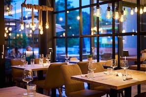 A restaurant interior with tables set for dining, hanging light fixtures, and large windows offering a view of the outside.