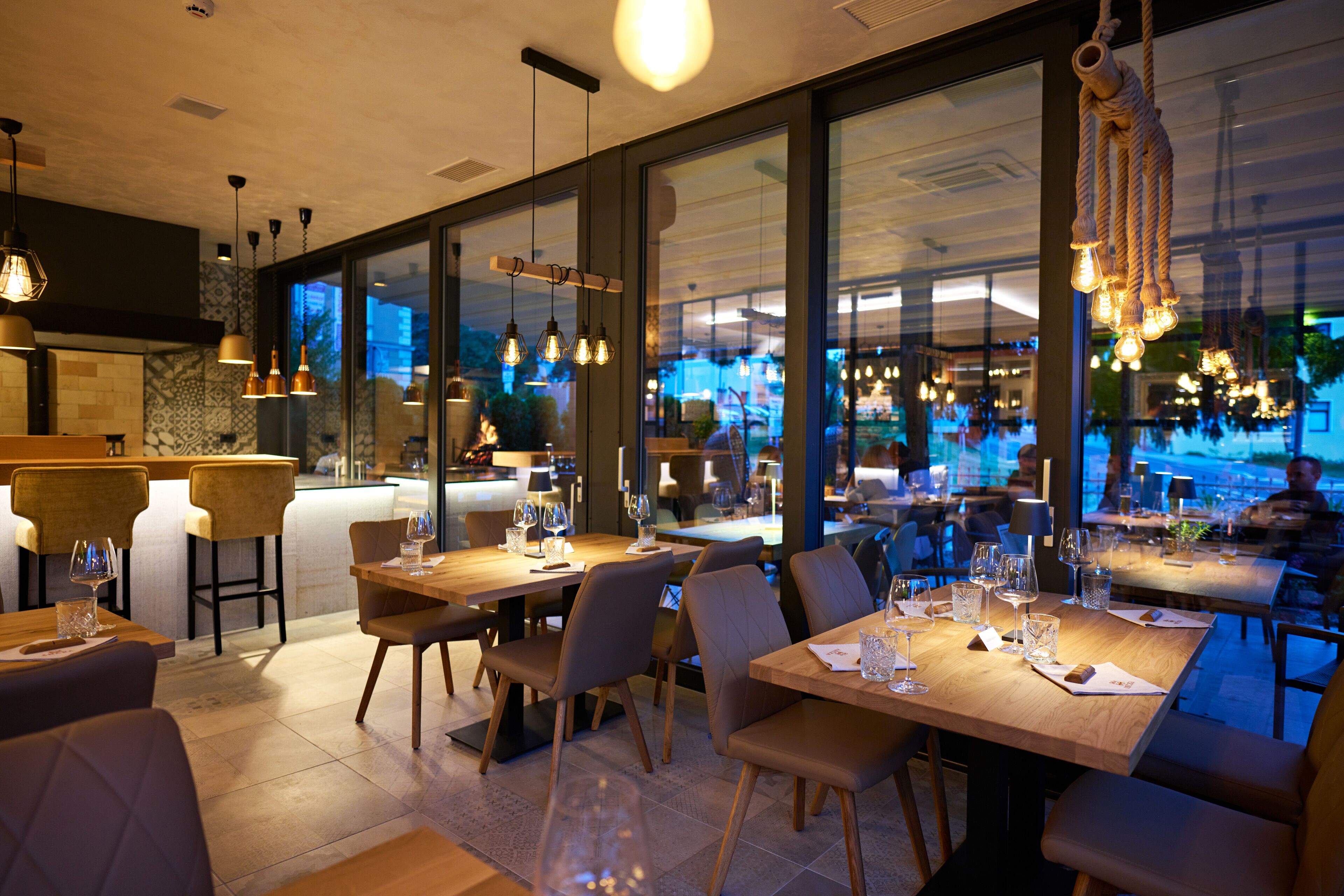 Ein Restaurant mit Holztischen und -stühlen, hängenden Leuchten und großen Fenstern.