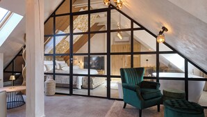 A loft bedroom with a sloped ceiling, a large window, a green armchair, a tufted ottoman, and a visible bathroom. - Hiša Ančka, Boutique Hotel (Slovenj Gradec)