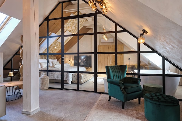 A loft bedroom with a sloped ceiling, a large window, a green armchair, a tufted ottoman, and a visible bathroom.