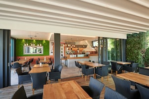 Un restaurante moderno con mesas de madera y sillas de mimbre negro, una pared verde y una cocina visible a través del vidrio.