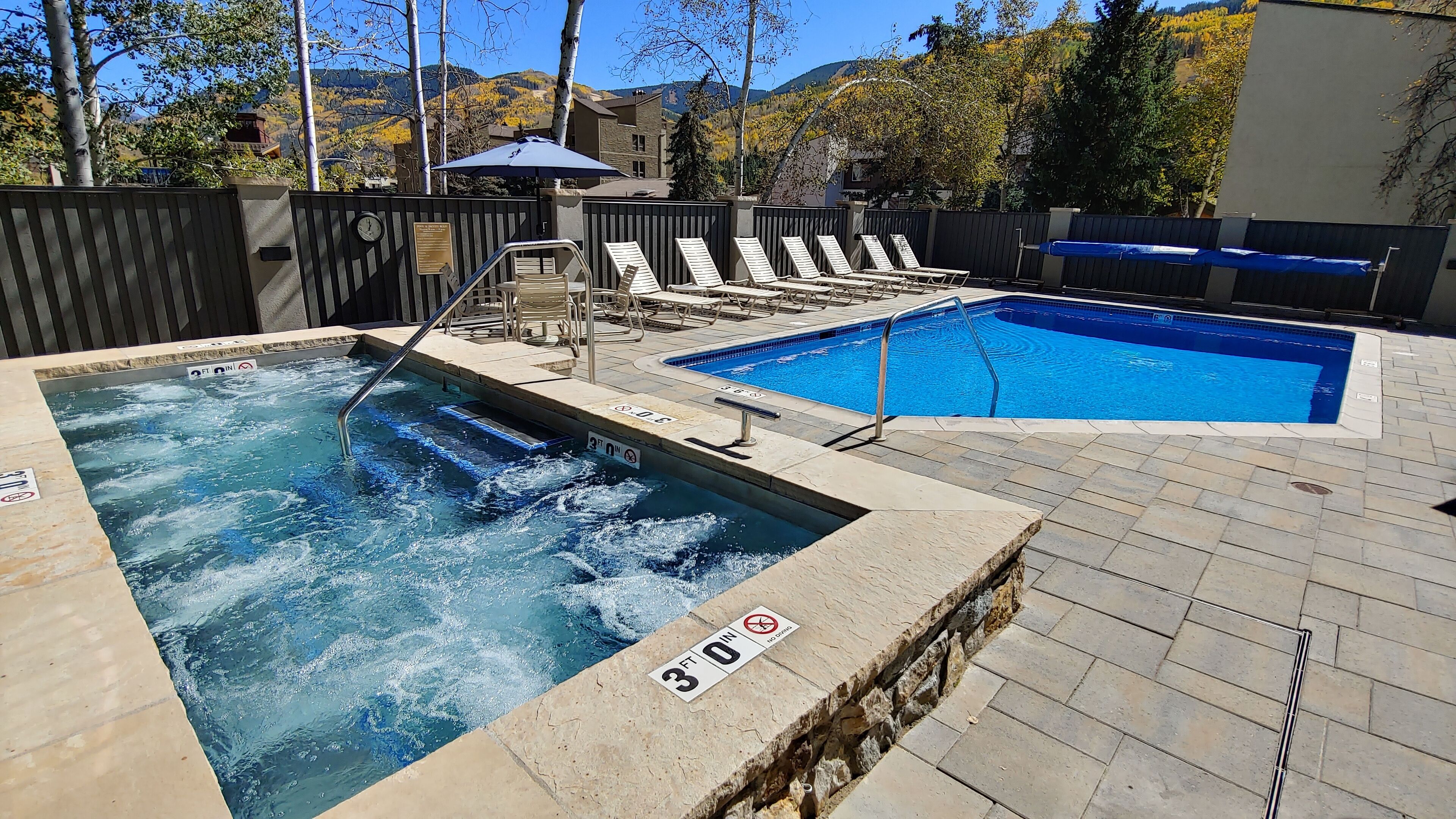 A hot tub and a swimming pool with lounge chairs, surrounded by a wooden fence and a mountain backdrop.