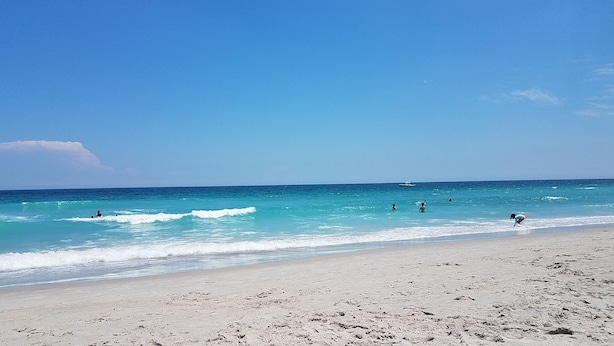 A beach with clear turquoise water, people swimming and walking along the shore, and a boat in the distance.