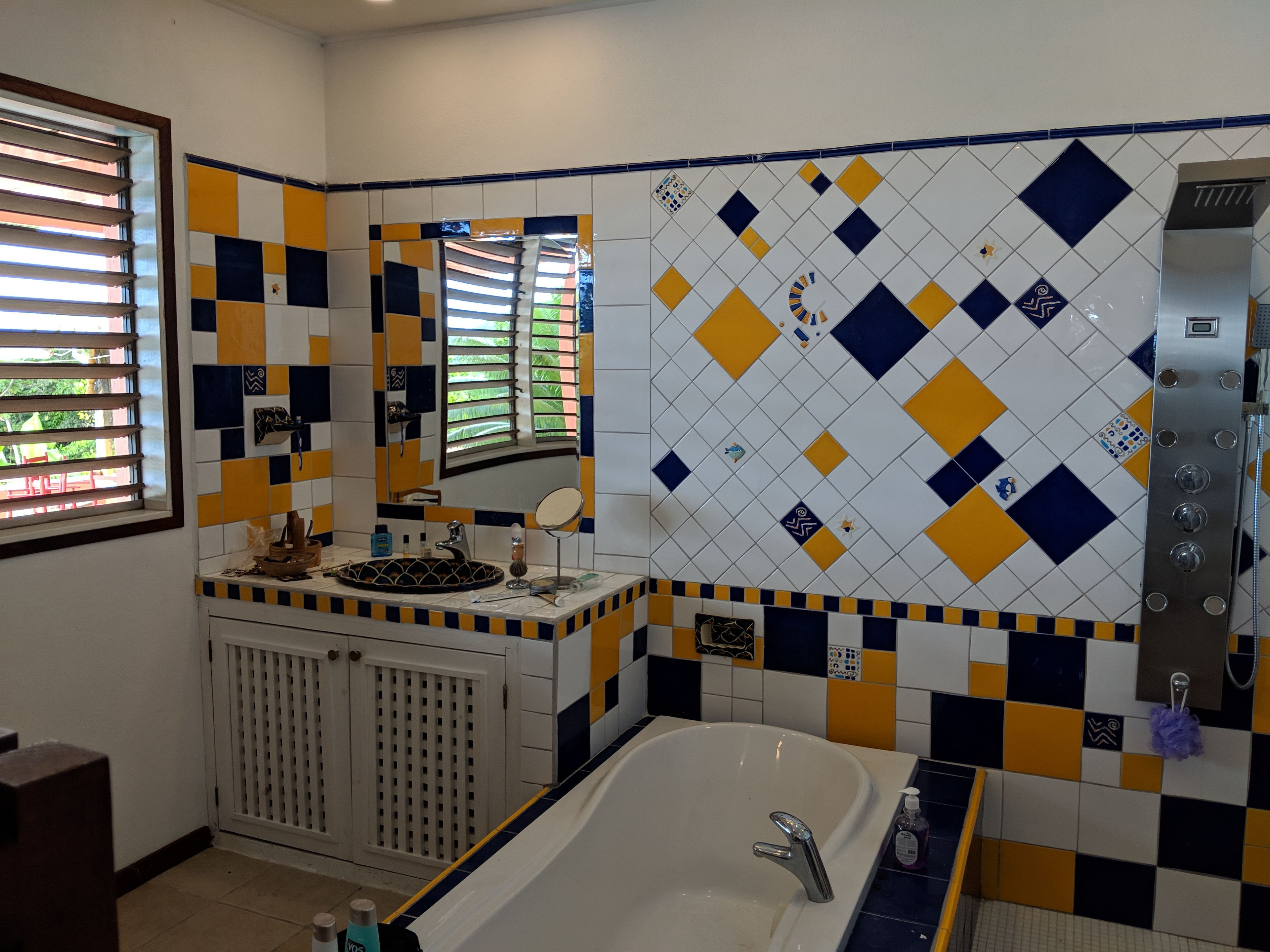 A bathroom with a bathtub, a sink, and a mirror. There is a window with blinds, and the walls are tiled with a diamond pattern.