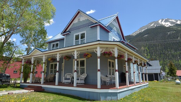 A two-story house with a porch, multiple windows, and a mountain backdrop. - Beautifully restored Victorian Mountain home furnished with authentic antiques! (Silverton)