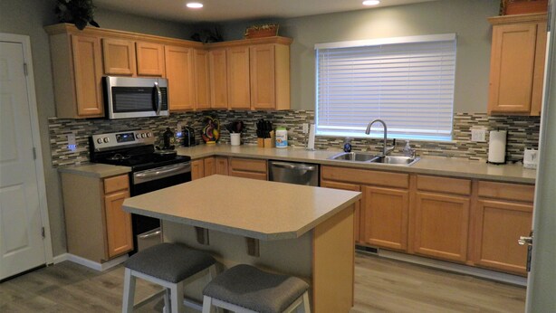 A modern kitchen with wooden cabinets, a stainless steel sink, a microwave, and a stove. There are two bar stools and a window with blinds. - Upscale Puyallup Home ☆ Near Seattle, Tacoma, Puget Sound, Cascade Mountains (Puyallup)