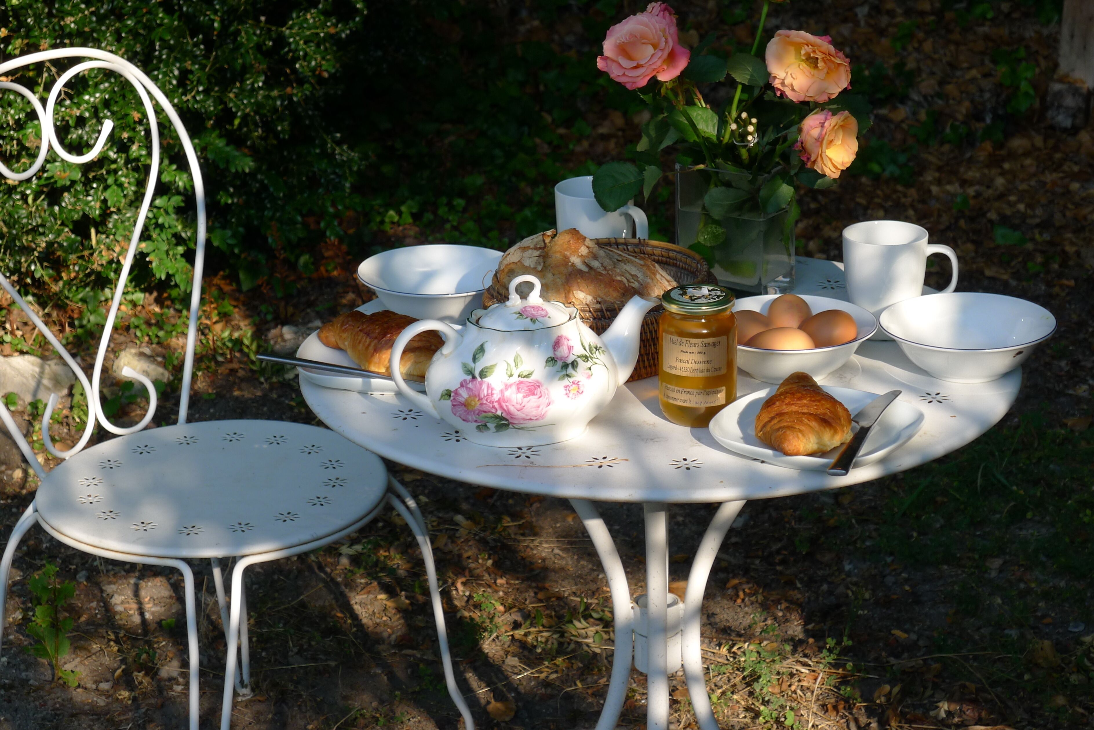 Een ronde witte tafel met een bloemendecor potje, kopjes, een mand met brood, een pot jam en een croissant.
