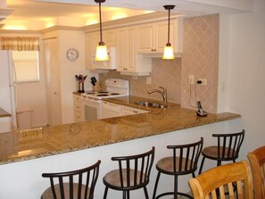 A kitchen with white cabinets, a granite countertop, a stove, and a sink. There are four bar stools and a wooden chair.