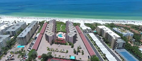 An aerial view of a hotel with multiple buildings, a central pool, tennis courts, and a beach in the background.