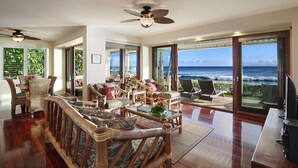 A spacious living area with a view of the ocean, furnished with wooden furniture and a ceiling fan. - Hale Nalu - Beachfront Home Steps from Baby Beach with Spa/AC! (Koloa)