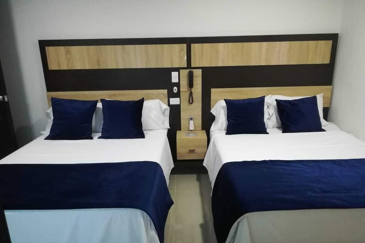 Ein Doppelzimmer mit zwei Betten, jeweils mit Holz Kopfteil, weißer Bettwäsche und blauen Kissen. Zwischen den Betten steht ein Nachttisch mit Telefon und einer kleinen Lampe.