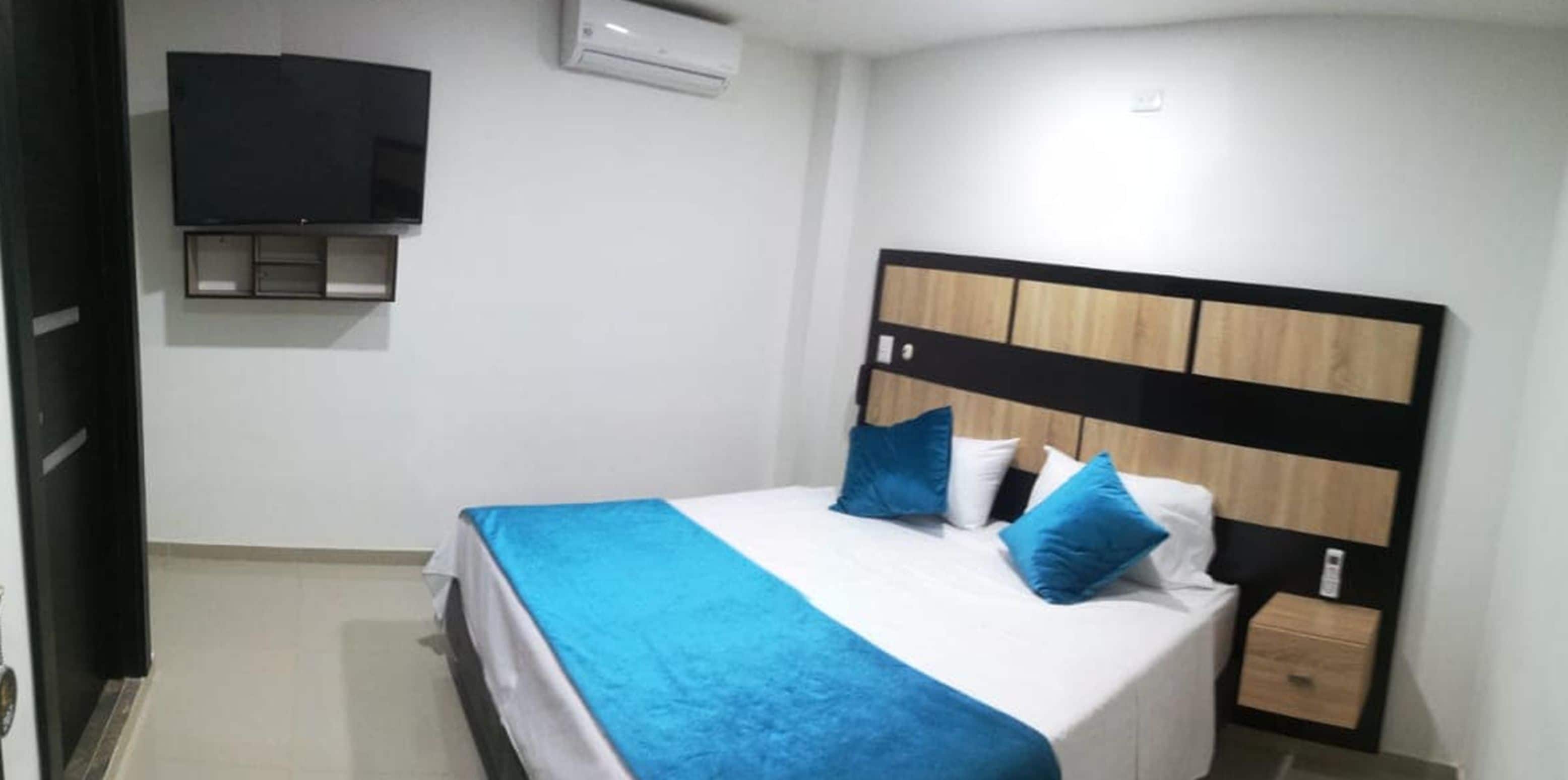 Hotellihuone, jossa on sänky, seinälle asennettu televisio ja pieni yöpöytä.