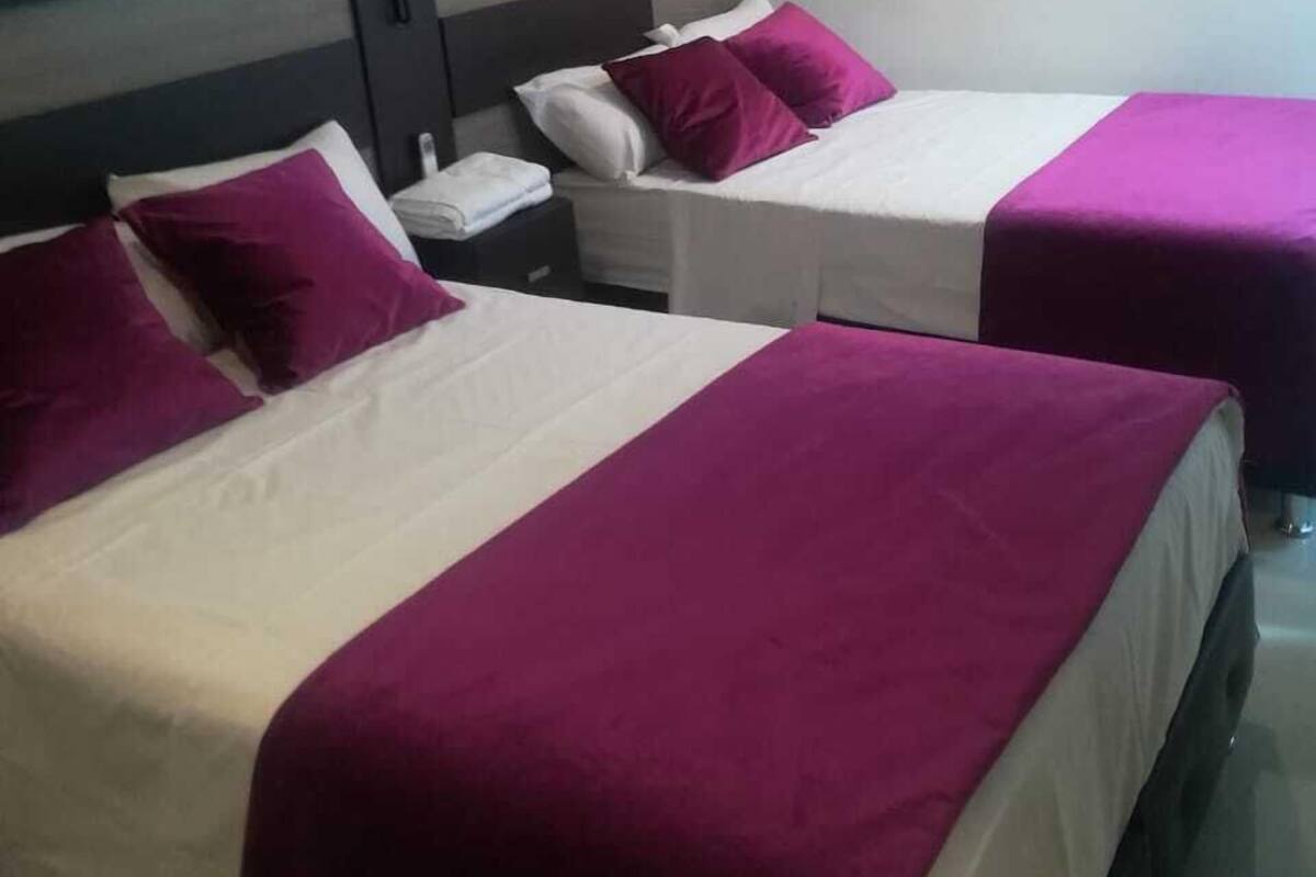 Ein Hotelzimmer mit zwei Betten, jeweils mit einer weißen und violetten Tagesdecke, einem grauen Kopfteil und einem Nachttisch mit einer Lampe.