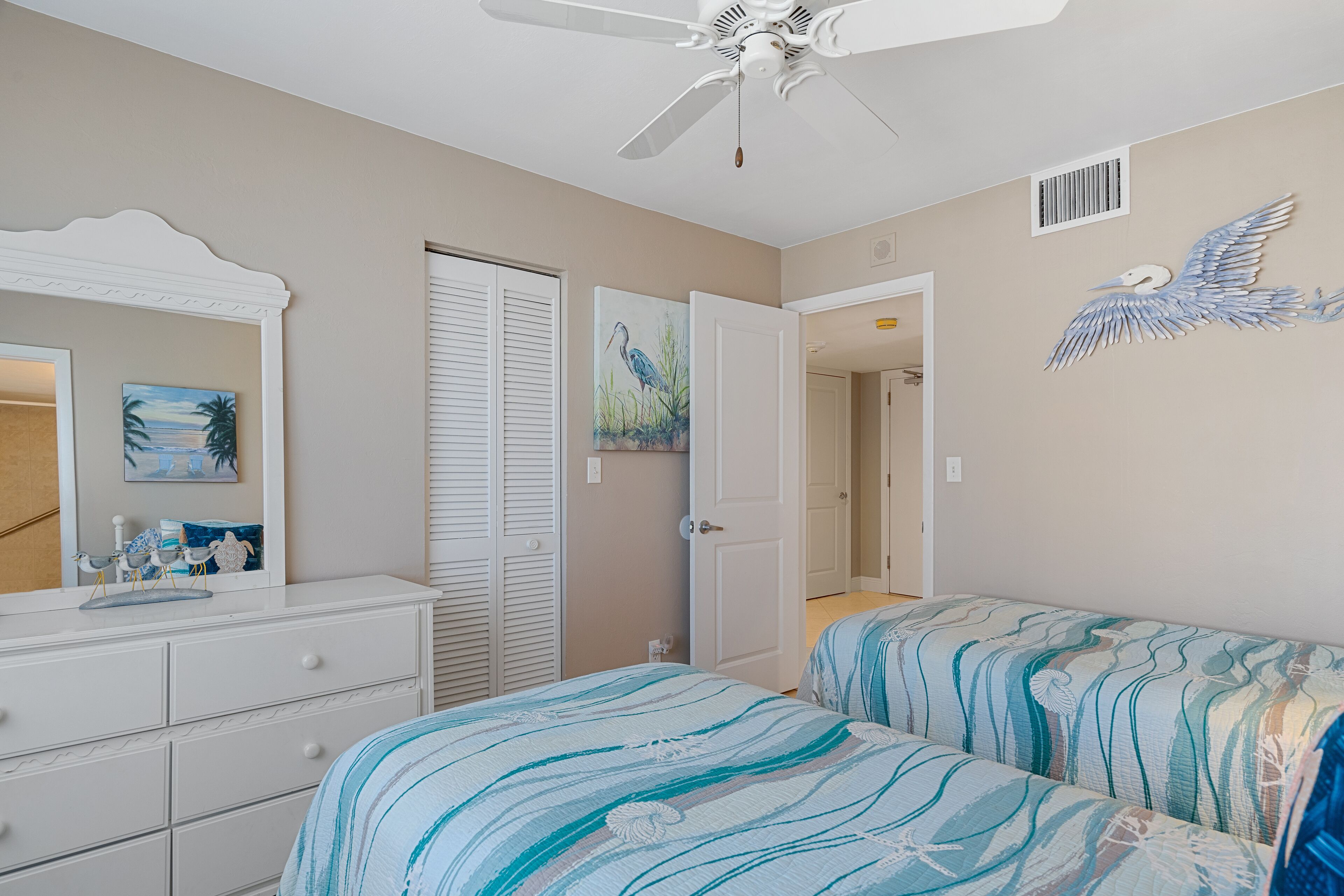 A bedroom with two beds, a dresser, a ceiling fan, and a door leading to another room.