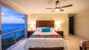A bedroom with a bed, two bedside lamps, a ceiling fan, and a view of the ocean.
