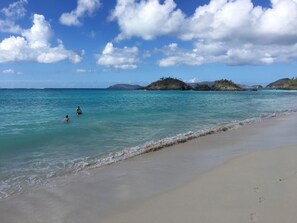 Una playa con agua turquesa cristalina, una orilla arenosa y unas islas rocosas en la distancia bajo un cielo parcialmente nublado.