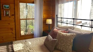 Un dormitorio de cabaña de madera con cama, almohadas, una mesita de noche, una lámpara y un ventanal con cortinas.
