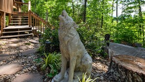 Una estatua de piedra de un lobo en un área boscosa, con una escalera de madera y un edificio rústico al fondo.