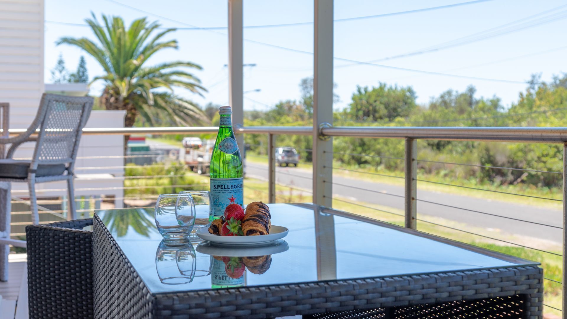 A glass table with a bottle of S.Pellegrini wine, a plate with food, and empty glasses on a balcony overlooking a road with palm trees and a car.