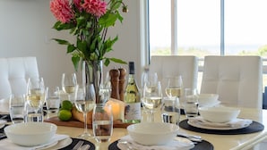 A dining table set with white plates, bowls, and wine glasses, a bottle of wine, a vase with flowers, and a loaf of bread. - Marilyn's - 180 degree views of Jervis Bay (Callala Beach)
