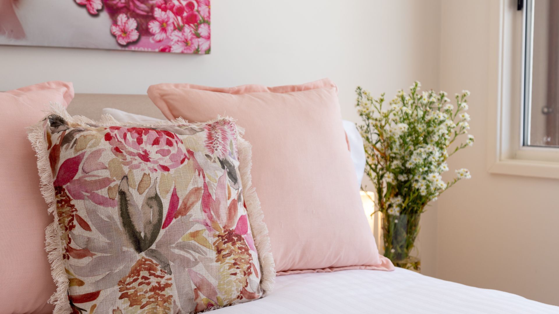 Une chambre à coucher avec un lit, deux oreillers, un coussin à motifs floraux et un vase contenant des fleurs blanches.